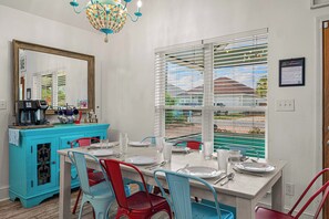 A dining area with a white table set for four, blue and red chairs, a turquoise cabinet with kitchen appliances, and a large window with blinds. - Family beach house w/ pool: 30A/Pier Park, Dog friendly, Golf Cart, West End PCB (Panama City Beach)