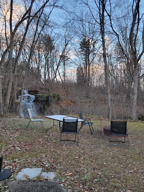 An outdoor seating area with a table and chairs, surrounded by trees and a small waterfall. - Rustic, Cozy Cottage with Backyard Waterfall and Cayuga Lake Views! (Ithaca)