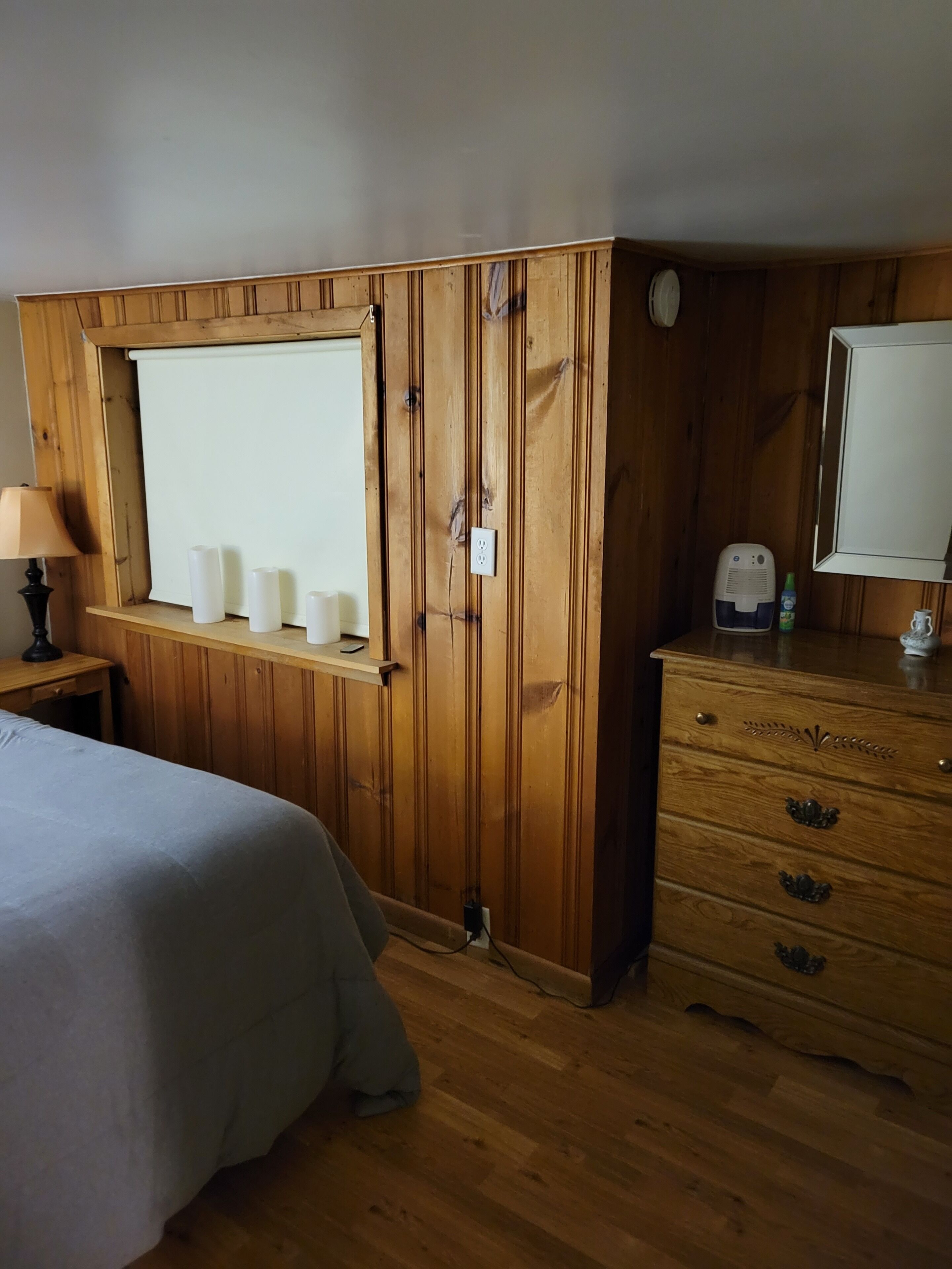 A wooden-paneled room with a bed, a dresser, a window with candles, and a mirror.