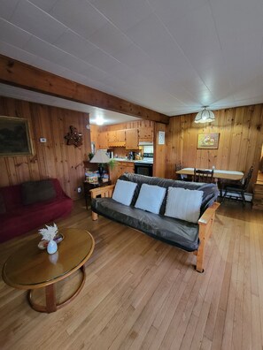 Een kamer met houten lambrisering, een bank, een eettafel en een keukenhoek.