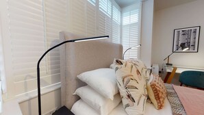 Un dormitorio con cama, almohadas blancas, una colcha estampada, un escritorio con lámpara y un cuadro en la pared.