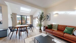 A modern living room with a brown leather sofa, a dining area with wooden tables and black chairs, and a large window with a view of a garden and a building.