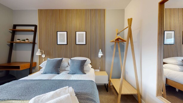 Una habitación de hotel moderna con una cama, mesitas de noche, un revestimiento de pared de listones de madera y un perchero de madera colgante.