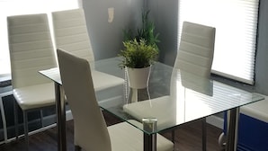 Un salón de conferencias con una mesa de vidrio, cuatro sillas blancas, una planta en maceta y ventanas amplias con persianas.