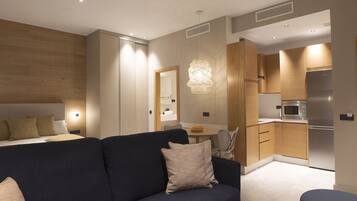 A modern hotel room with a dark blue sofa, a small round table, and a kitchenette with a stainless steel refrigerator and microwave.