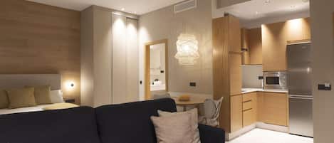 Une chambre d’hôtel moderne avec un canapé bleu foncé, une petite table ronde et une kitchenette équipée d’un réfrigérateur et d’un four à micro-ondes en acier inoxydable.
