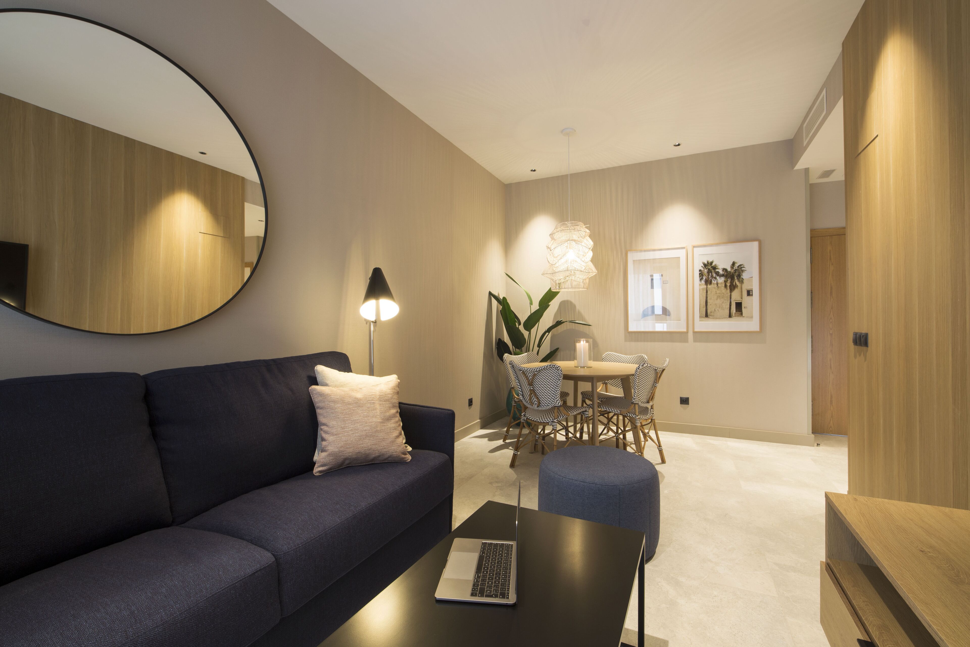 A modern living room with a dark blue sofa, a round mirror, a table with a laptop, a dining area with a table and chairs, and framed pictures on the wall.