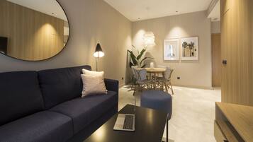 Een moderne woonkamer met een donkerblauwe bank, een ronde spiegel, een tafel met een laptop, een zithoek met een tafel en stoelen, en ingelijste foto’s aan de muur.