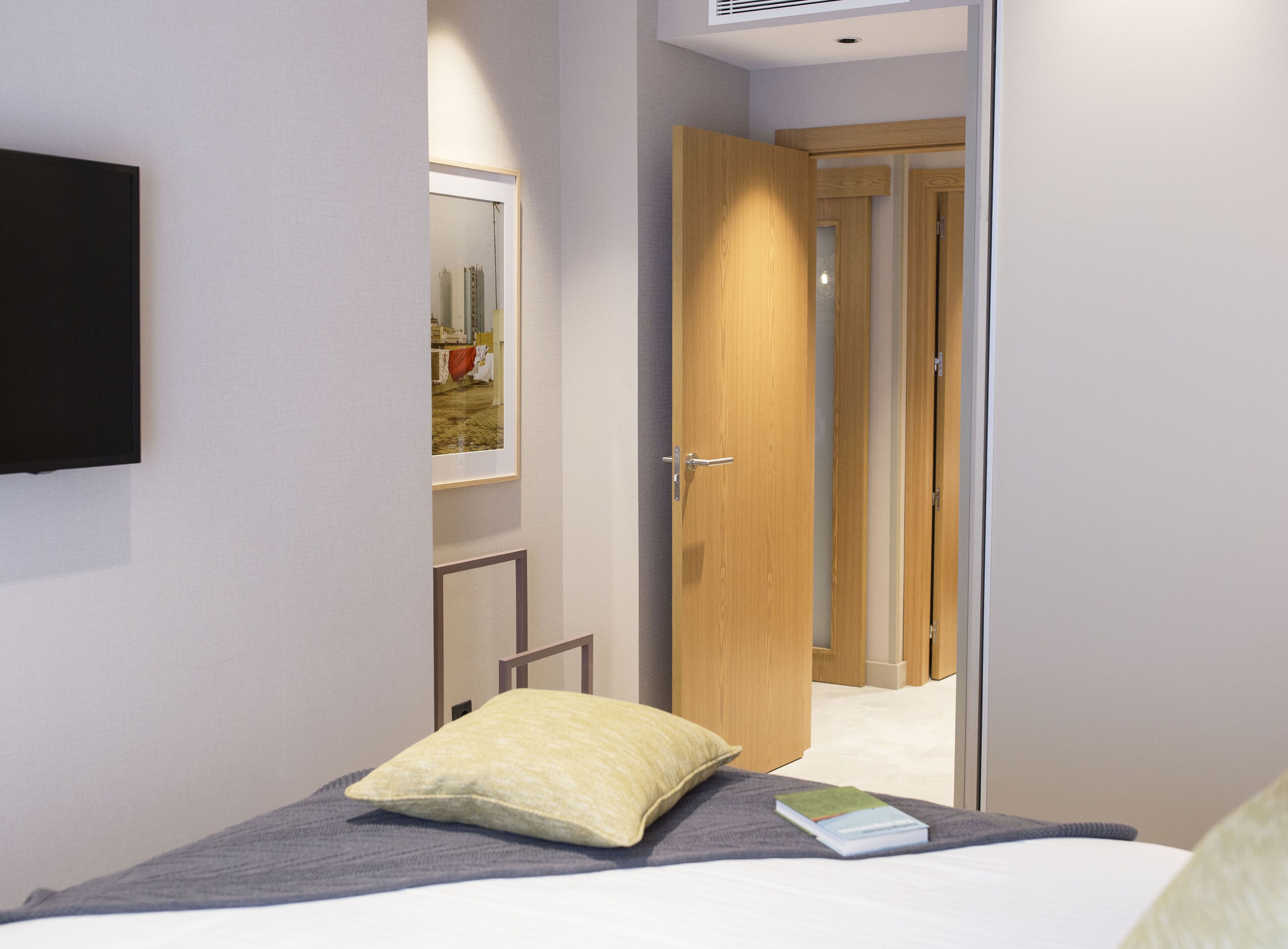 Un dormitorio con una cama, una mesita de noche, un almohadón y un libro. Se ve una puerta que da a otra habitación.