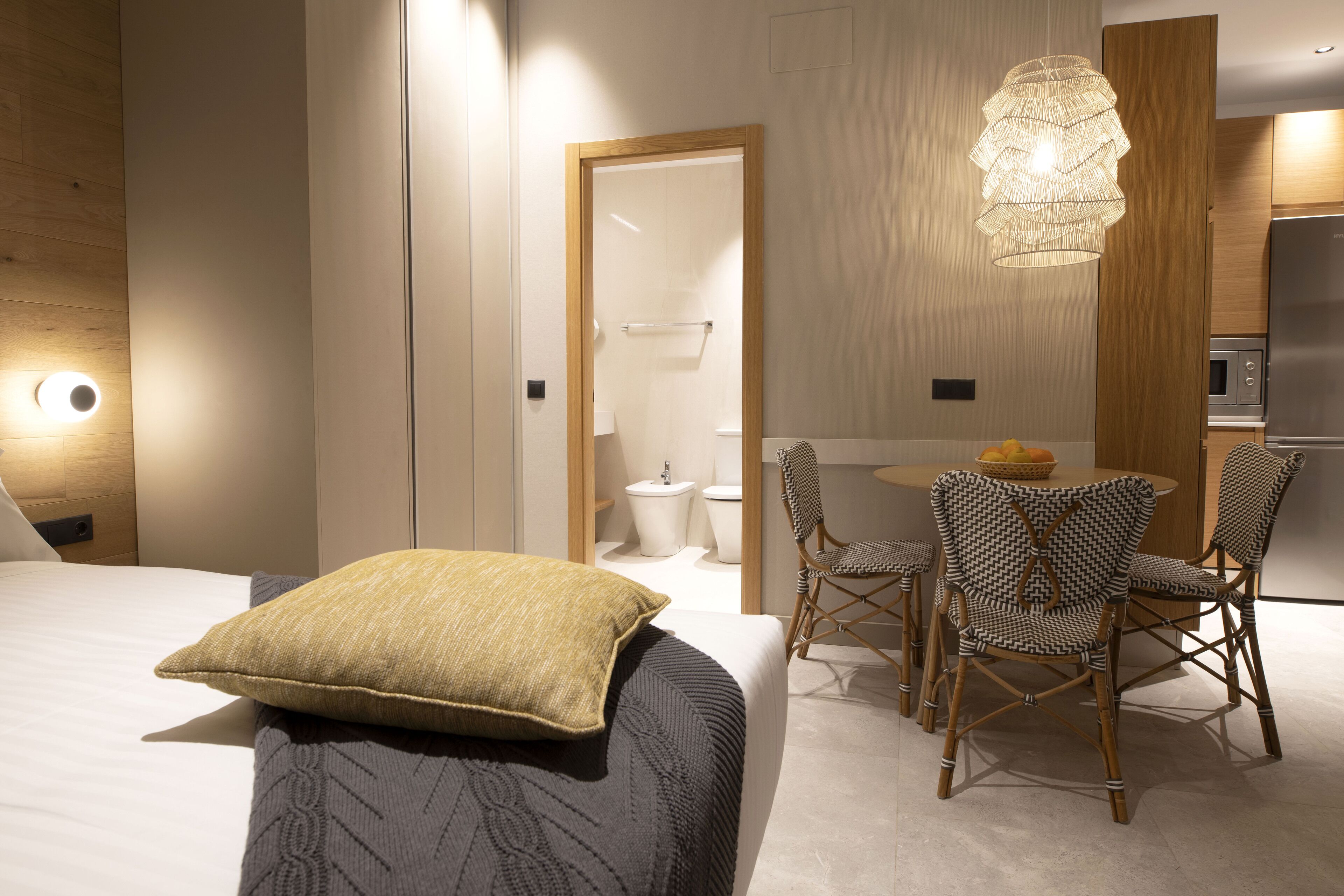 A modern hotel room with a bed, a dining table with chairs, a kitchenette, and a bathroom visible through a glass door.