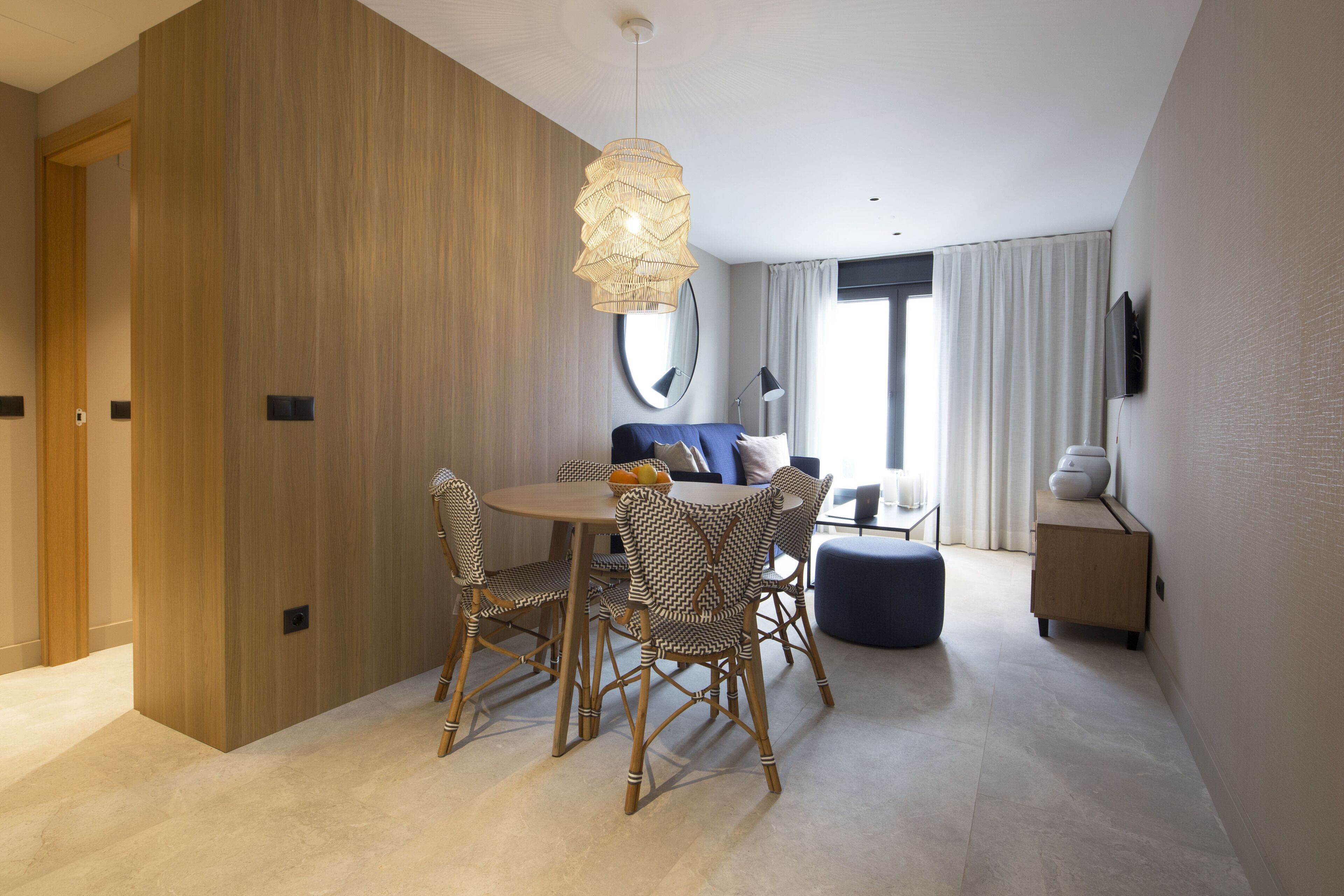 A modern hotel room with a dining area featuring wicker chairs, a round table, and a hanging light fixture.