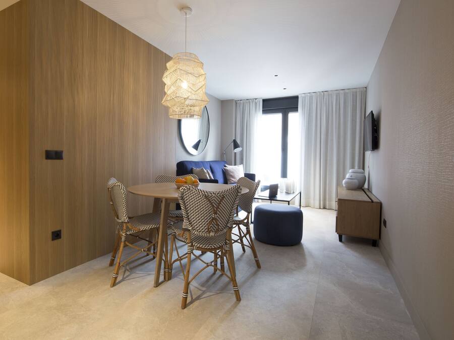 A modern hotel room with a dining area featuring wicker chairs, a round table, and a hanging light fixture.