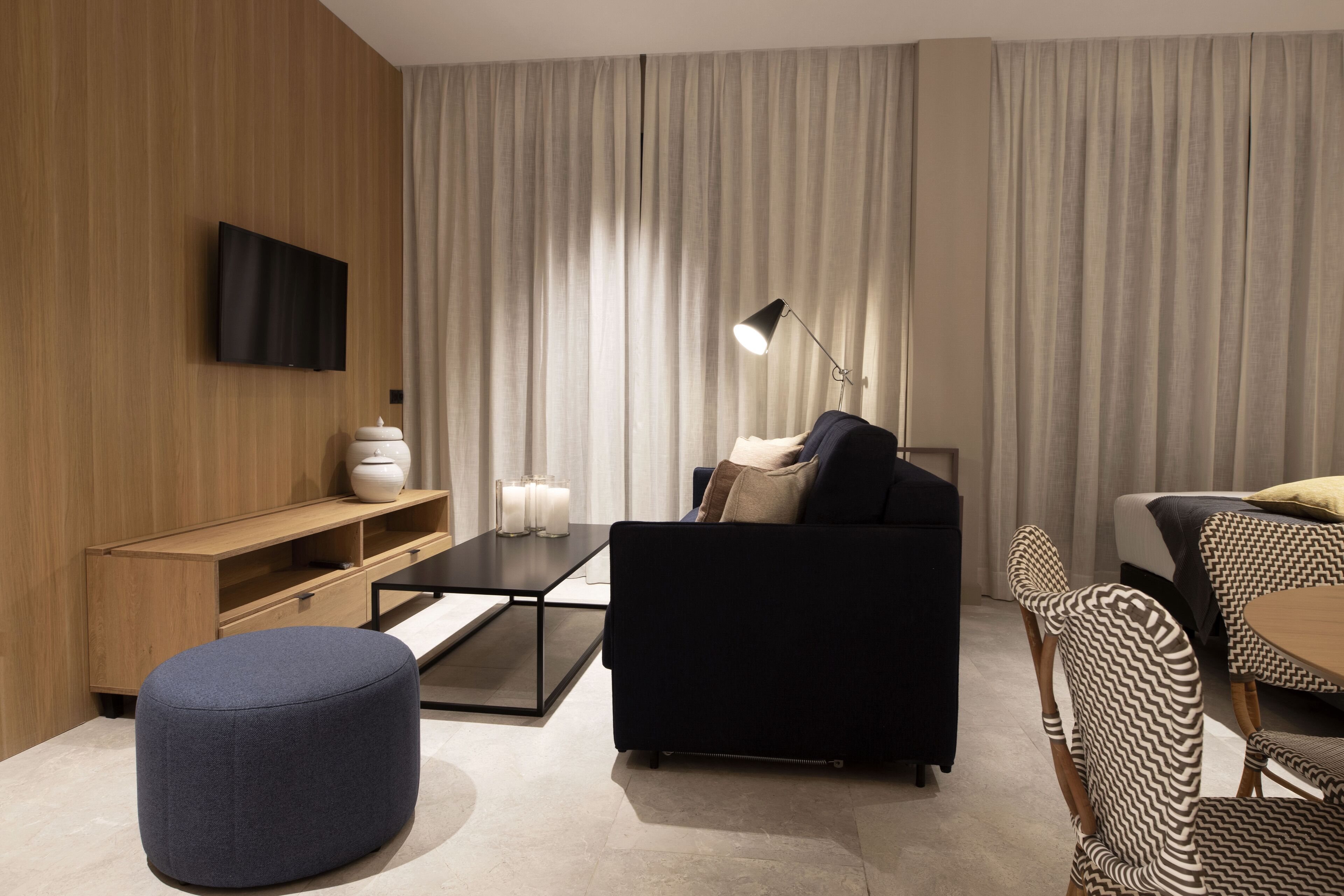 A modern living room with a flat-screen TV mounted on a wooden wall, a black sofa with cushions, a small round ottoman, a glass coffee table, and a floor lamp.