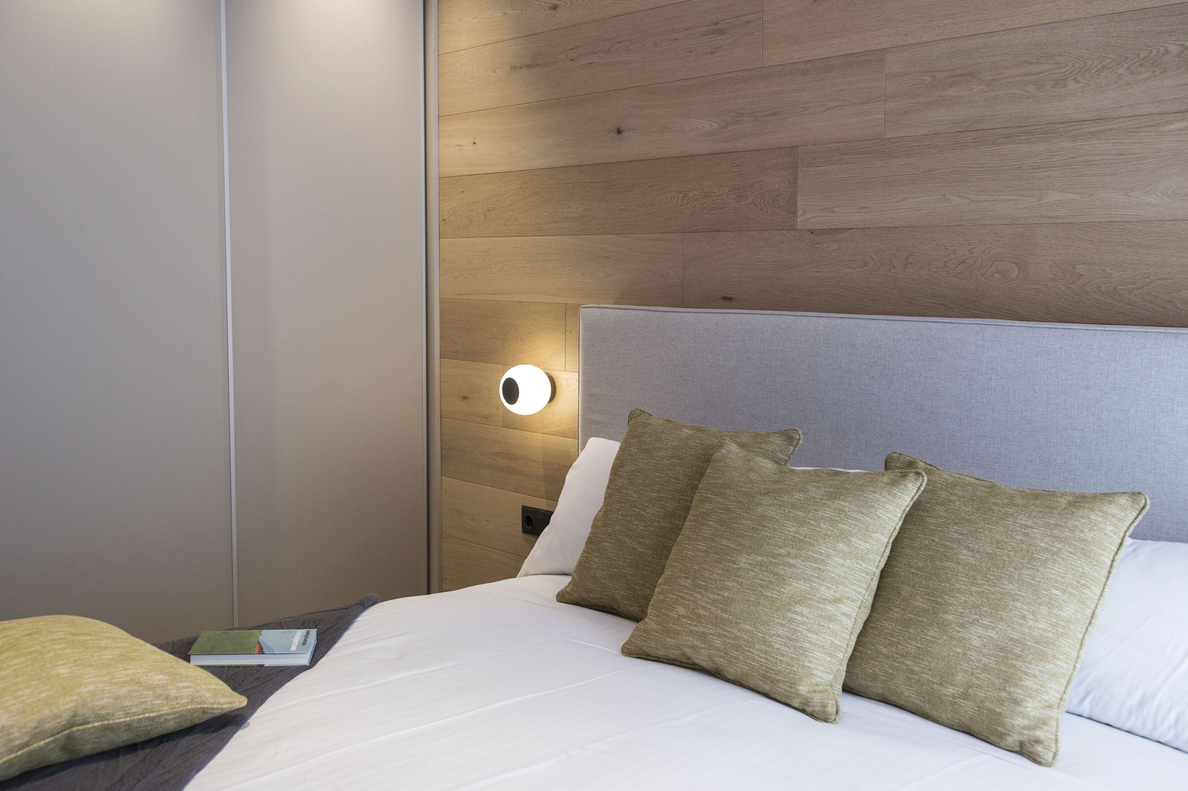 Una cama bien hecha con sábanas blancas y dos almohadas, una mesita de noche con lámpara y un panel de madera en la pared.