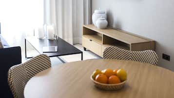 A modern dining area with a round wooden table, wicker chairs, a bowl of fruit, a television on the wall, and a window with curtains.