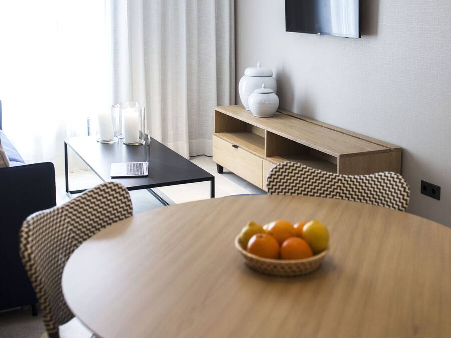Un comedor moderno con una mesa redonda de madera, sillas de mimbre, un cuenco de fruta, un televisor en la pared y una ventana con cortinas.