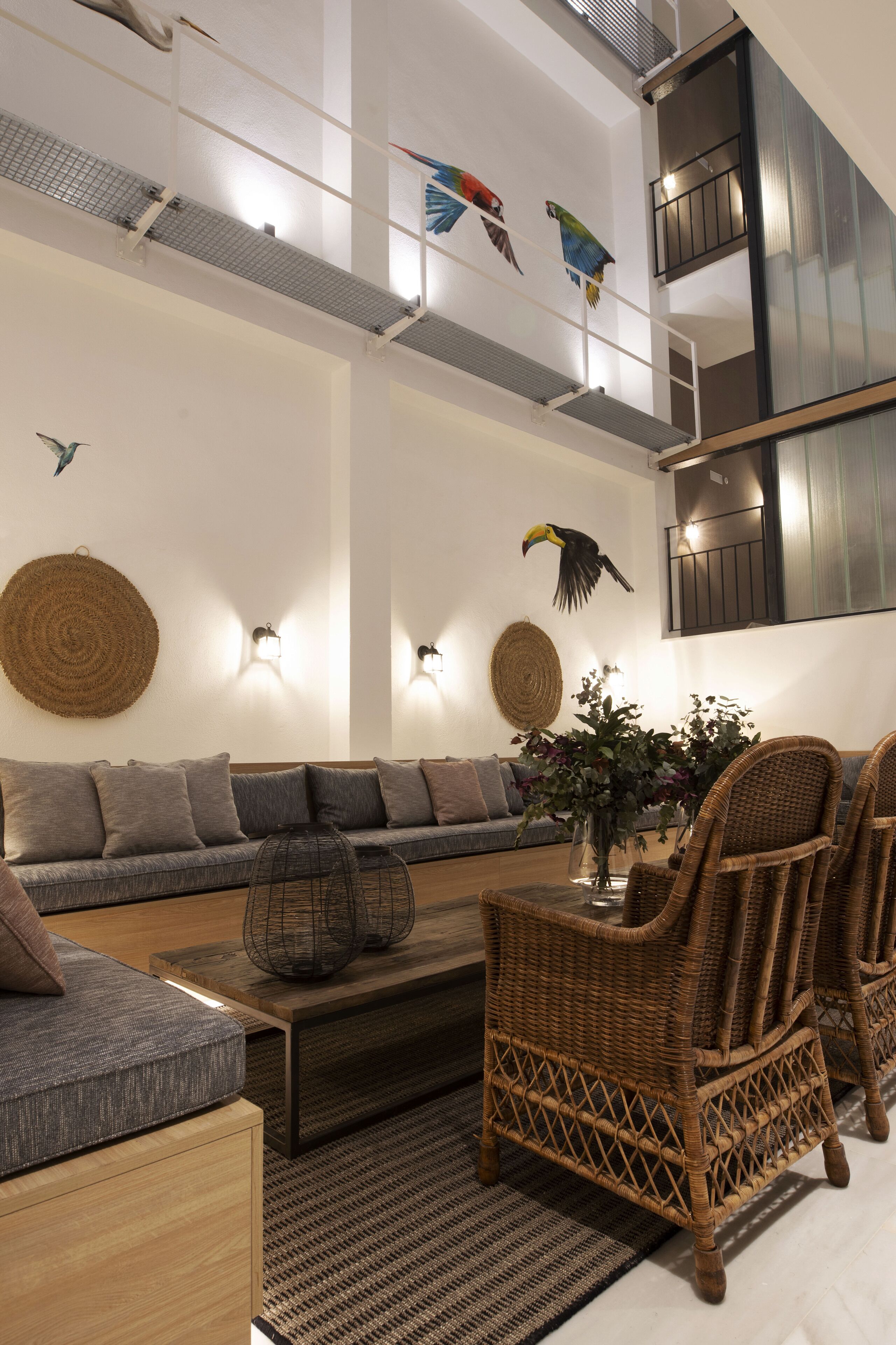 A modern living room with a grey sofa, wicker chairs, and a coffee table. Decorative wall hangings and wall-mounted lights are visible.
