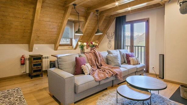 Uma sala de estar aconchegante no estilo loft, com teto alto, vigas de madeira, um sofá cinza com almofadas, uma mesa redonda pequena e vista para montanhas através de grandes janelas.