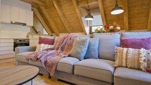 Uma sala de estar aconchegante em estilo loft, com teto alto, vigas de madeira, um sofá com almofadas, uma mesa de centro redonda e uma área de cozinha com armários e fogão.