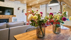 Uma área de refeições com uma mesa de madeira, dois vasos de vidro com flores, um sofá cinza e uma TV fixada na parede.