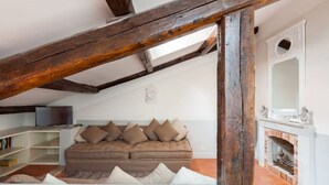 A loft-style room with exposed wooden beams, a sofa with cushions, a fireplace, and a television on a shelf.