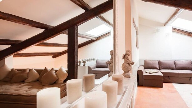 A cozy loft bedroom with a sloped ceiling, exposed wooden beams, a large window, a plush sofa, and a row of candles.