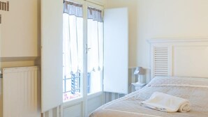 A bedroom with a large bed, a window with curtains, a radiator, and a nightstand with a lamp.