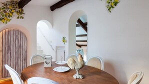 A round wooden dining table with white chairs, a decorative rooster centerpiece, and a rustic wall with an arched doorway.
