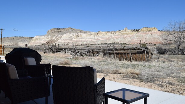 A patio with wicker chairs and a table, overlooking a rocky landscape with a wooden fence and a small building.