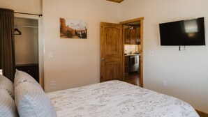 A bedroom with a bed, a wooden ceiling, a painting, a TV, and a kitchen in the background.