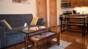 A living room with a gray sofa, wooden coffee table, and a kitchen area with wooden cabinets and appliances.