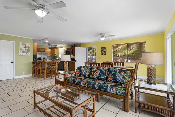 A spacious living area with a sofa, coffee table, and dining table. There is a kitchen in the background with cabinets and a refrigerator.