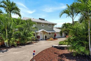 A two-story house with a front porch, surrounded by tropical plants and a gravel driveway.