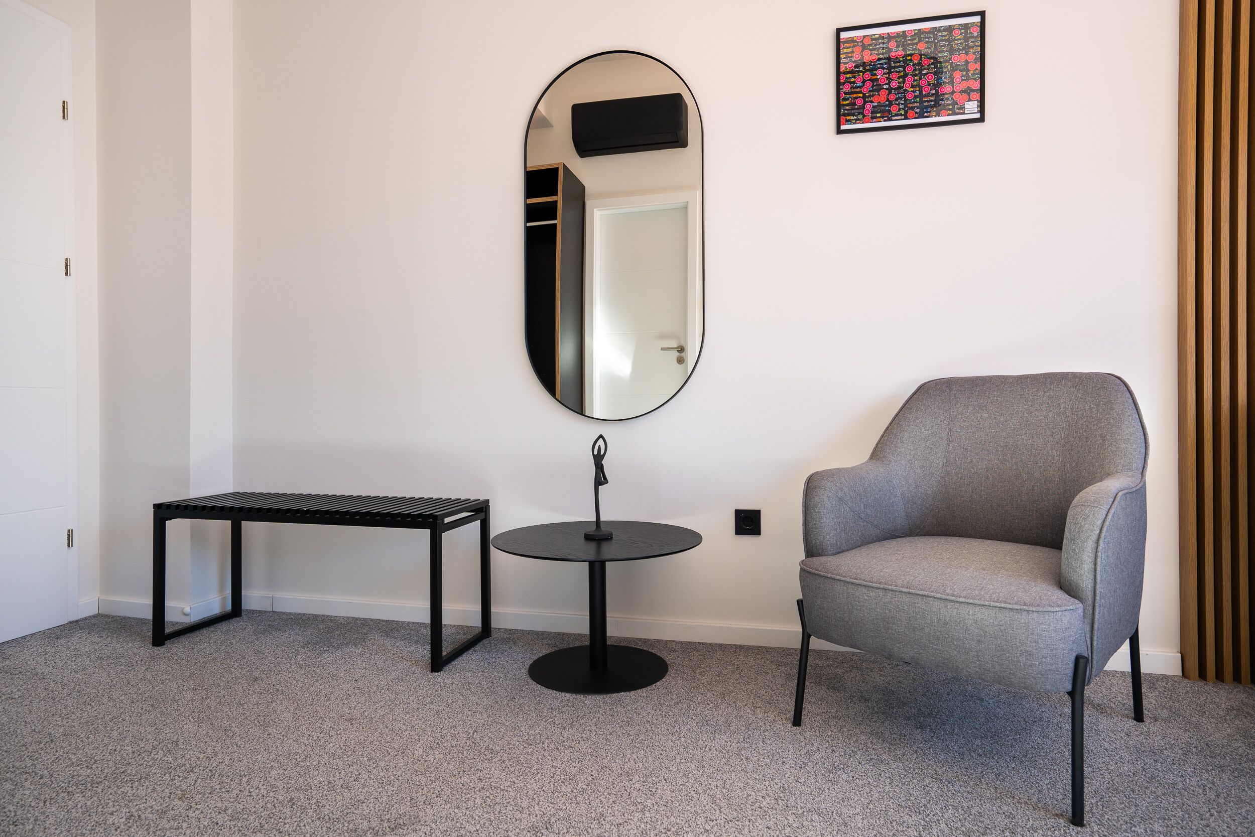 Una habitación con un sillón gris, una mesita redonda pequeña, un banco y un espejo grande en la pared.