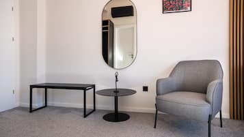 Una habitación con un sillón gris, una mesita redonda pequeña, un banco y un espejo grande en la pared.