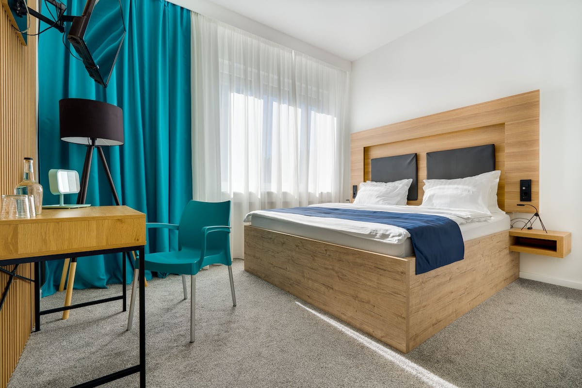 A hotel room with a wooden bed, a desk, a chair, a lamp, and a window with curtains.