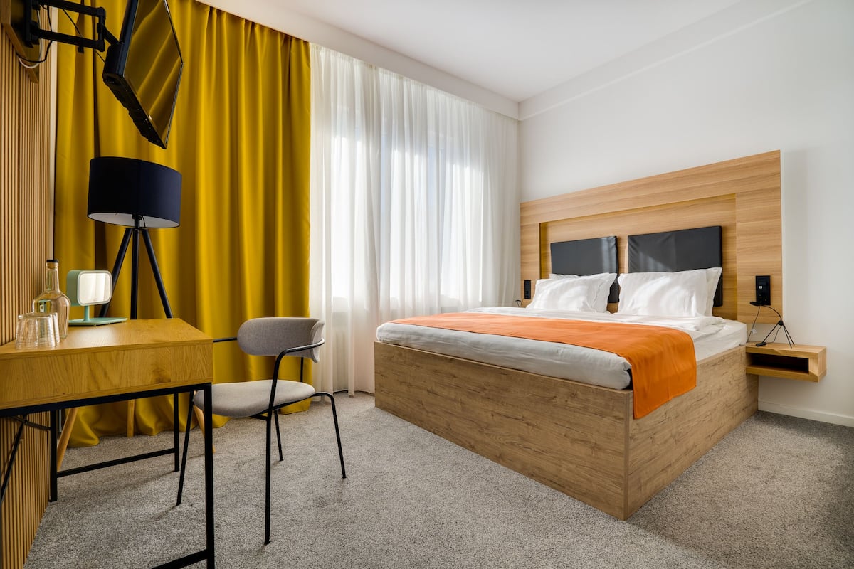 A hotel room with a wooden bed, a desk, a chair, a lamp, and a large window with yellow curtains.