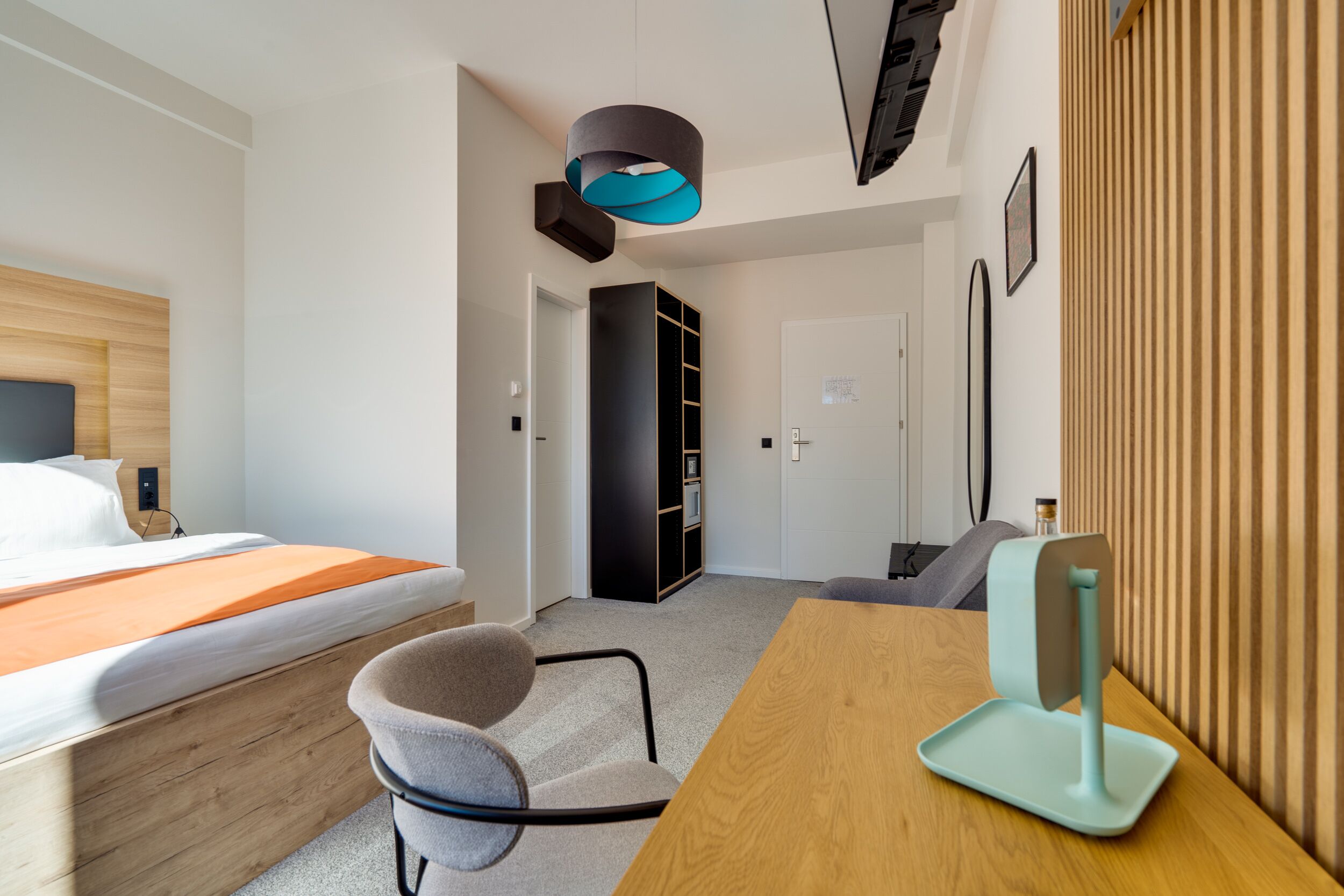 Une chambre d’hôtel moderne équipée d’un lit, d’un bureau, d’une chaise, d’une bibliothèque et d’un luminaire au plafond au design original.