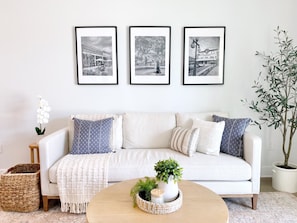 A living room with a white sofa, three framed black and white photos on the wall, a small potted plant, and a woven basket.
