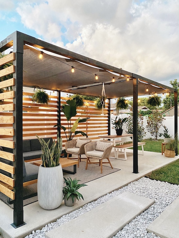 An outdoor patio area with a wooden pergola, string lights, wicker furniture, and potted plants.