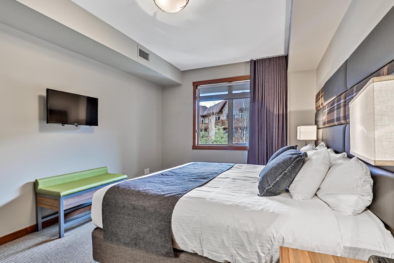 Quarto de hotel moderno com cama, um banco, televisão e janela com vista para casas.