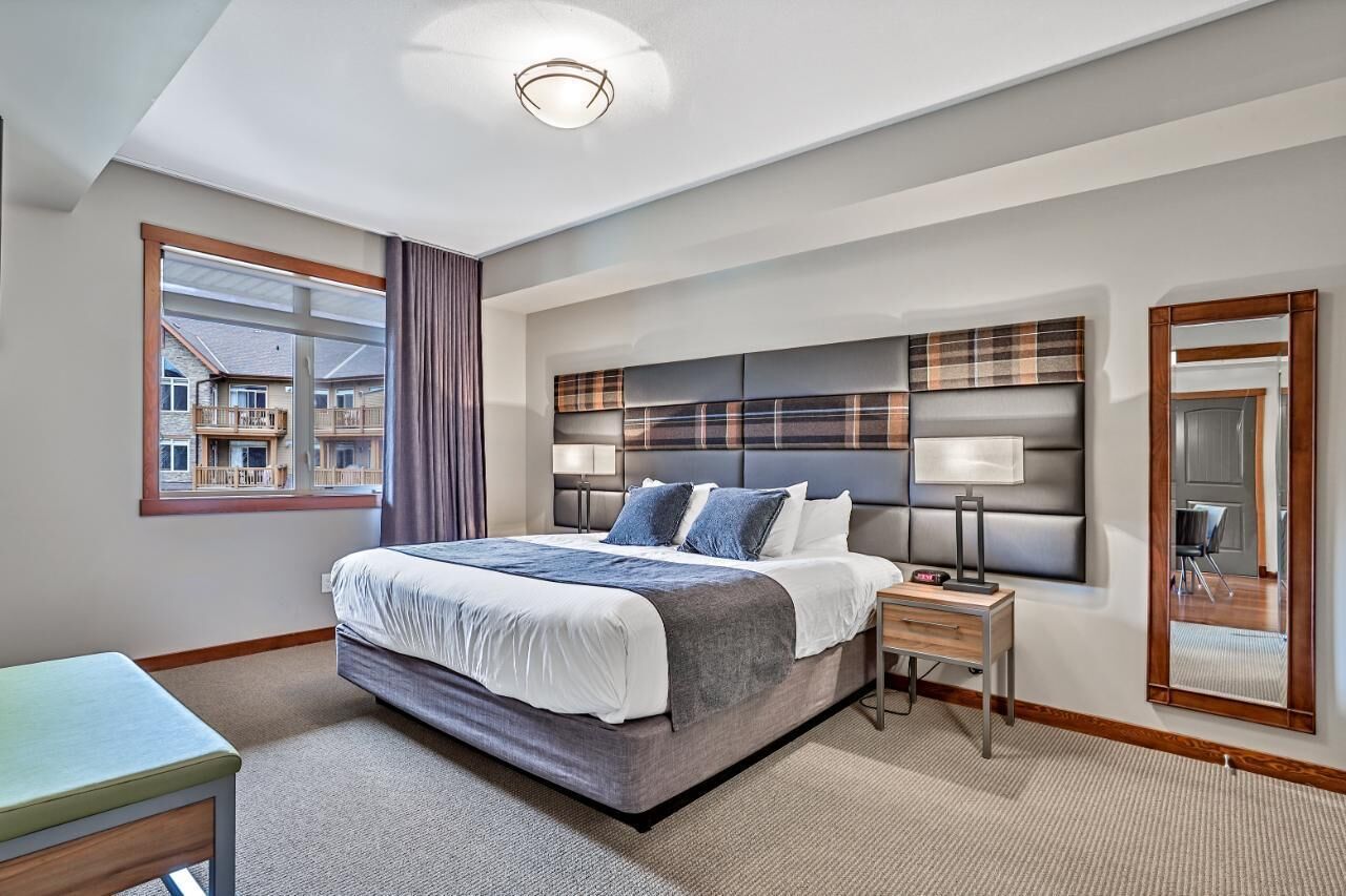 A modern hotel room with a large bed, a bedside table, a nightstand, a mirror, and a window with a view of other hotel buildings.