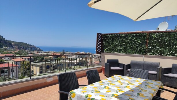 A terrace with a table and chairs, a large umbrella, and a view of the sea and buildings.
