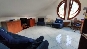 A spacious living area with a high ceiling, a television, a dining table with chairs, and a large window providing a view of the outdoors.