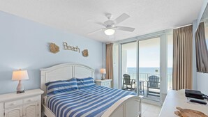 Una camera da letto con un letto, comodini, una ventola a soffitto, vista sulla spiaggia e un balcone con una sedia.