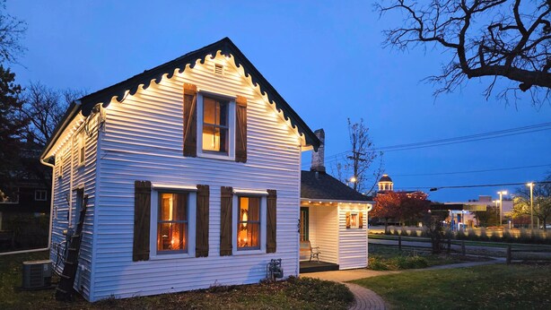 A white house with a porch and a chimney, surrounded by a grassy area and a brick path. - The Tailor House (Woodstock)
