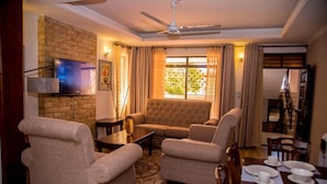 A living room with a sofa, armchairs, a TV mounted on a brick wall, a ceiling fan, and a dining area with a table set for a meal. - The Festival Hotel Suites & Spa (Mtwapa)