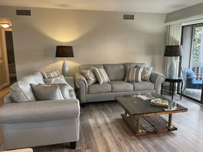 A living room with a grey sofa set, two side tables, a glass coffee table, and a view of the outdoors through a large window.
