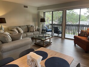 A spacious living room with a grey sofa, a coffee table, and a dining table with chairs. There is a sliding glass door leading to an outdoor patio area. - The Pinehurst Perch - Pool in complex (Pinehurst)