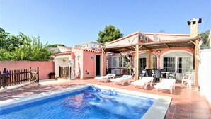 Una piscina con sillones de descanso, un porche techado y un edificio rosa con techo de tejas.